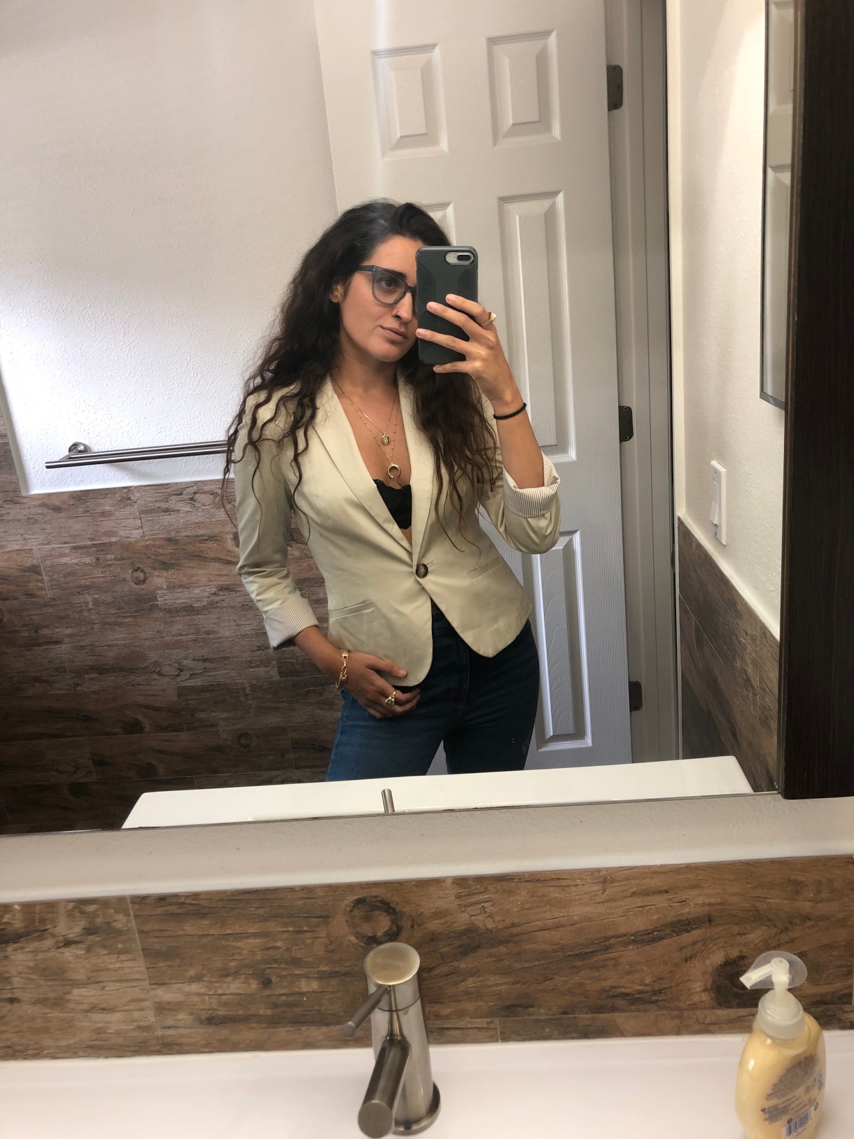 Kayla Kumari Upadhyaya wears a beige blazer buttoned over just. a black lacey bra and ripped jeans. She also wears two gold necklaces and is taking a selfie in the mirror.
