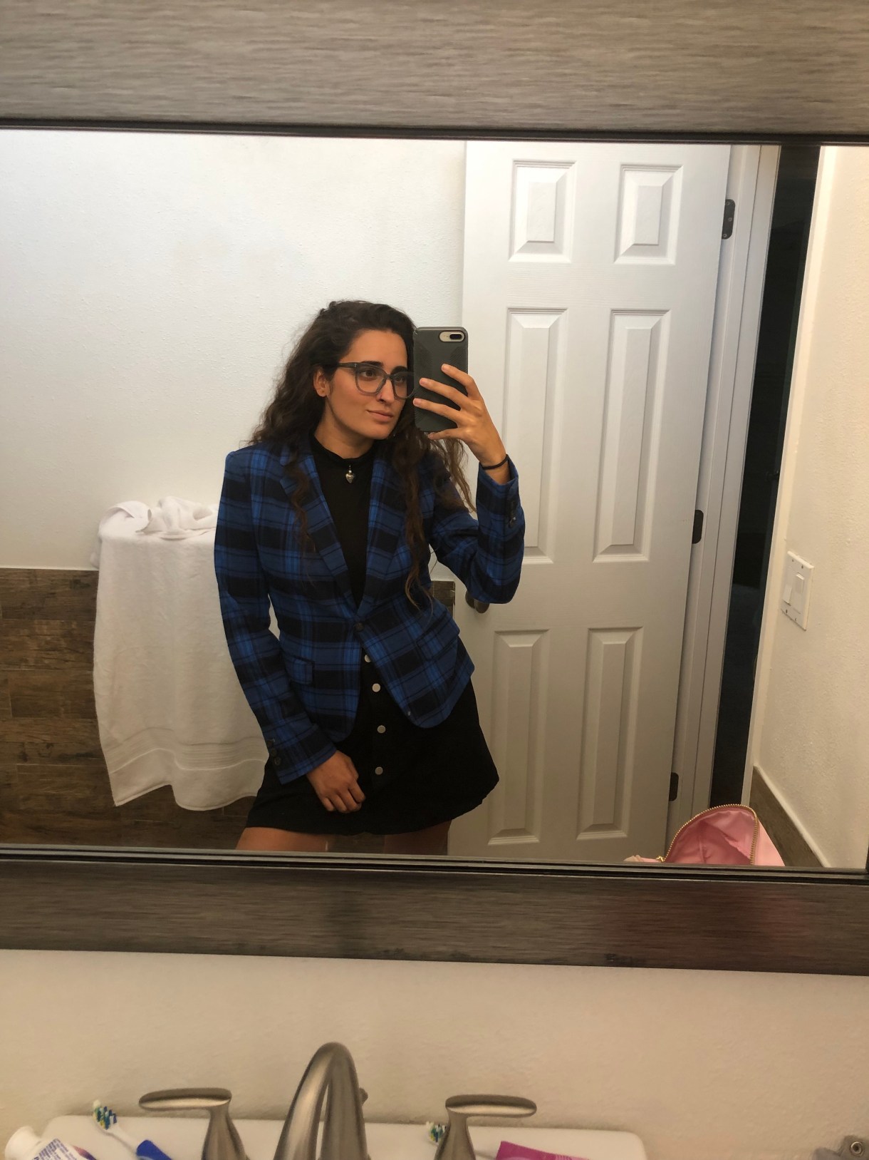 Kayla wears a black miniskirt and black turtleneck tank top with a blue and black plaid blazer that is buttoned. She's taking a mirror selfie.