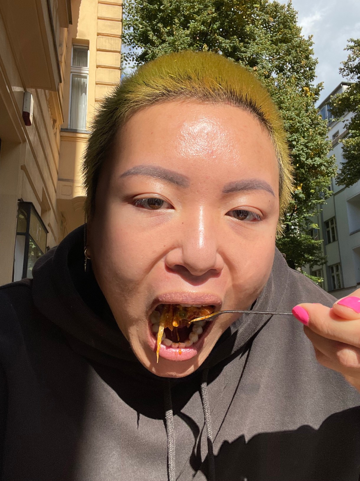 viv is an asian human here eating out of doors, their mouth super wide open. they have dyed green hair and pink finger nails and are wearing a hoodie