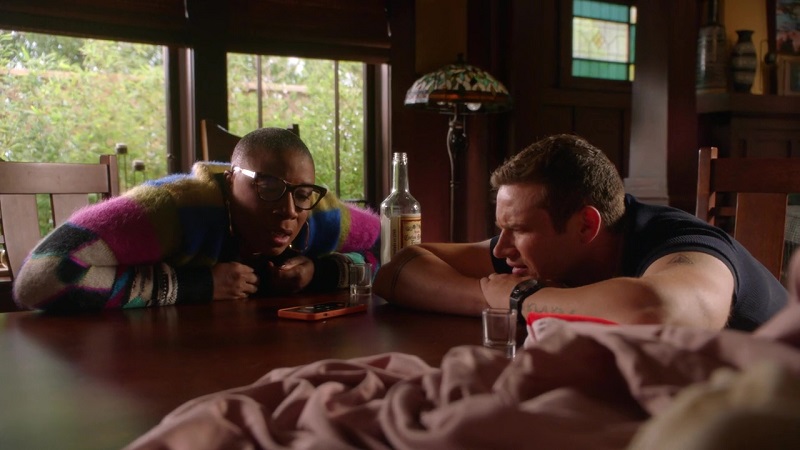 Hen and Buck sitting at the table and relaying instructions to their colleagues at the 118. Hen is wearing a Graphic-Pattern Knitted Jumper while Buck's wearing a black t-shirt. A half empty bottle of liquor stands between them.