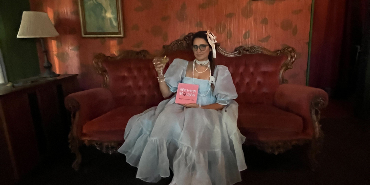 Kayla sits on an ornate Victorian sofa holding aloft a cocktail in a stemmed glass. She is wearing a doll-esque baby blue dress that is all gauzy with incredibly puffy sleeves, layers of pearls and a lace choker, ribbons in her hair which is braided, and glasses. She is holding her book, Helen House, which is pink with the title on it in white whimsical font and the