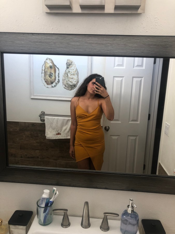 Kayla Kumari Upadhyaya wears an orange tight asymmetrical mini dress, and her long curly brown hair is down. An oyster painting is behind her.