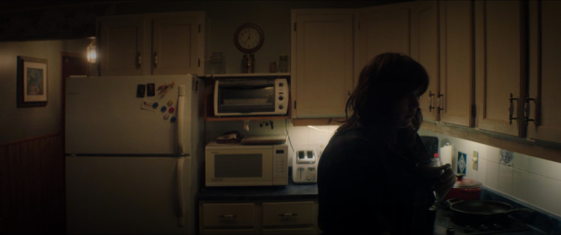 Teagan Johnston in shadows stands in a kitchen holding a wine glass. 