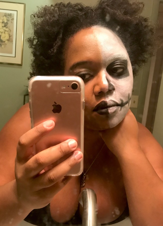 Carmen, a Black woman with tightly curled medium length black hair parted in the middle takes a selfie in the mirror with half her face in skull makeup and the other half in black lipstic and accompanying shimmery eyeshadow