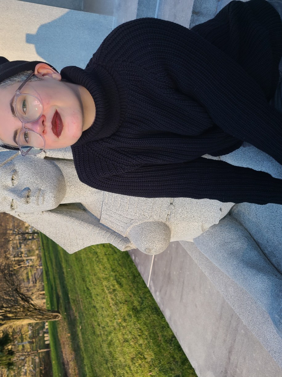 Nico, a white genderqueer human with blonde hair leans against a sphynx, wearing a black turtleneck and beanie as well as glasses and burgundy lipstick