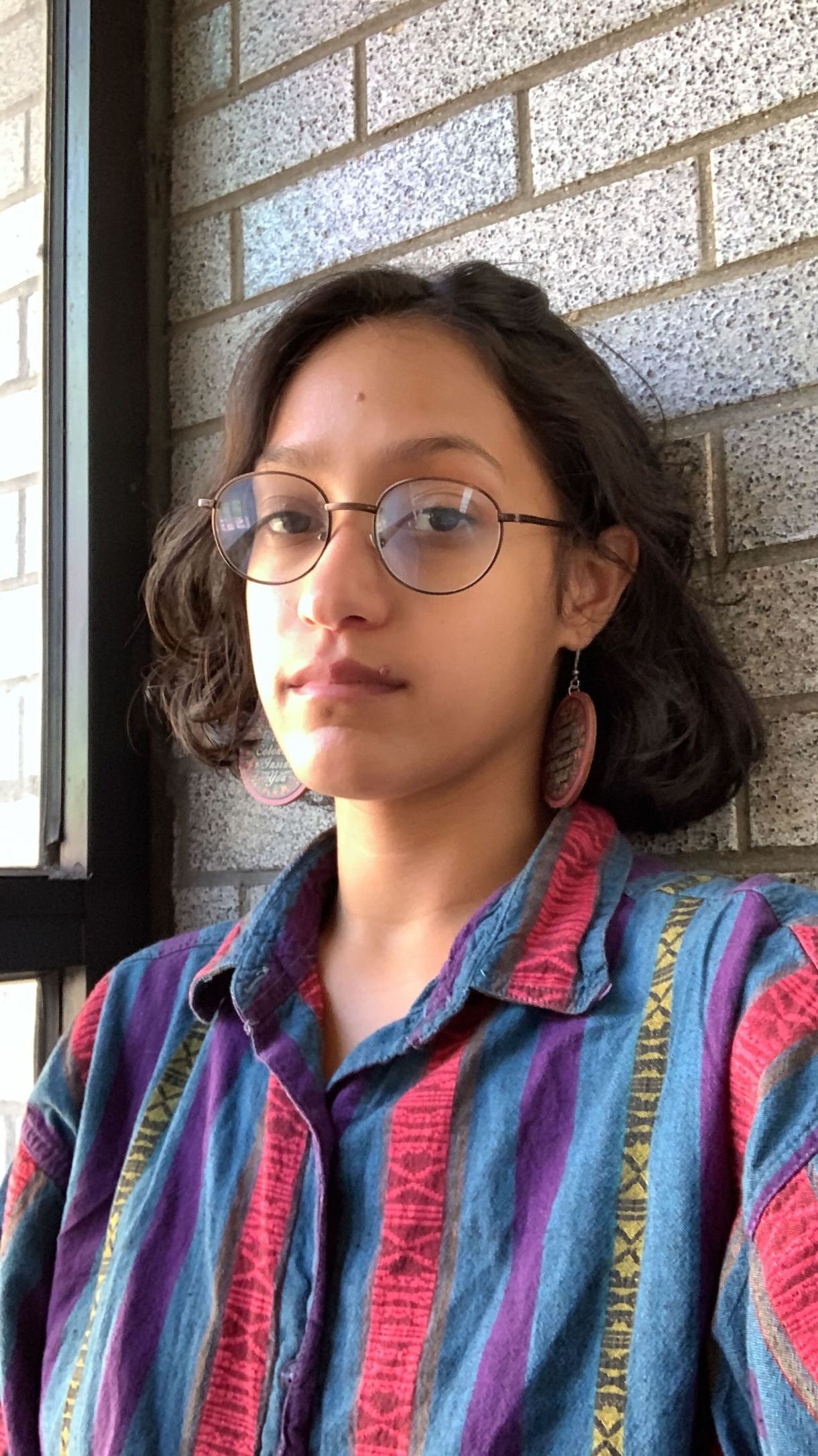 Lily, a Puerto Rican woman with neck-length brown hair and glasses, gazes at the camera. She is wearing a colorful button down shirt and earrings.