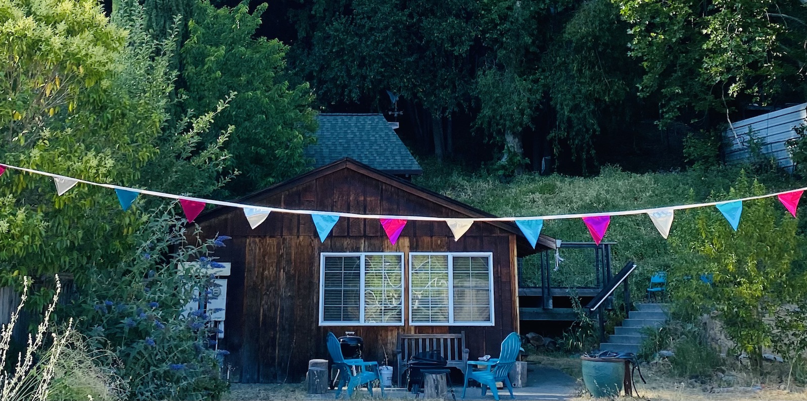 Mugworts Cabin: a cute cabin with a rainbow triangle flag banner in front sits in the woods, surrounded by tall lush green trees