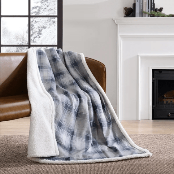 a blue and gray checked blanket over a brown couch against a white wall with an open window. on the white wall, there is a white fireplace