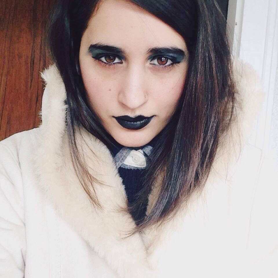 kayla, with straightened dark brown hair, looks at the camera wearing black lipstick, heavy eyeliner and blue eyeshadow, and a fluffy furry white coat