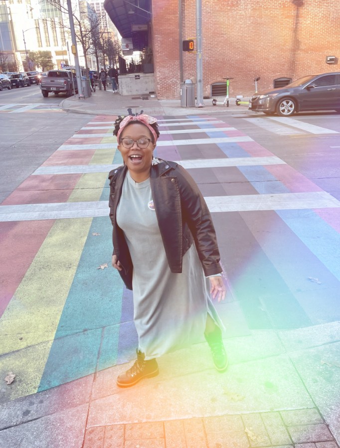 Carmen, a Black femme, is in a green long dress with a Black leather motorcycle jacket and her hair bulled up in a pink scarf. She has on combat boots. She's standing outside on a street corner with where the street is painted rainbow and also there is a rainbow sun glare on the camera.