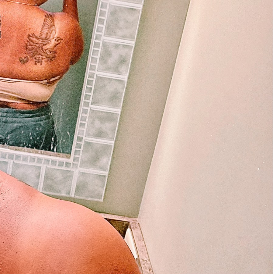 A picture of Carmen's back in a splotchy mirror, showing off her tattoo. Carmen has brown skin. The tattoo is a black outline of a bird with large wings and two small black images of a Taino symbol of the Coqui frog and the Sankofa symbol.
