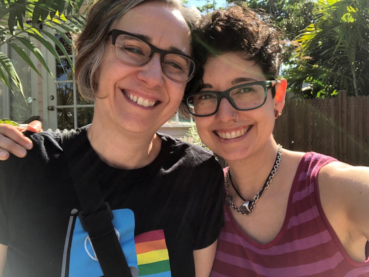 Mia and Tracy stand in a sunny, palm-dense outdoor area in Key West. This photo looks fairly recent. Mia has her short, brown streaked with gray, bob, and Tracy's hair is short and culry on top. They both wear glasses. Tracy jas a mpse ring and Mia an eyebrow ring