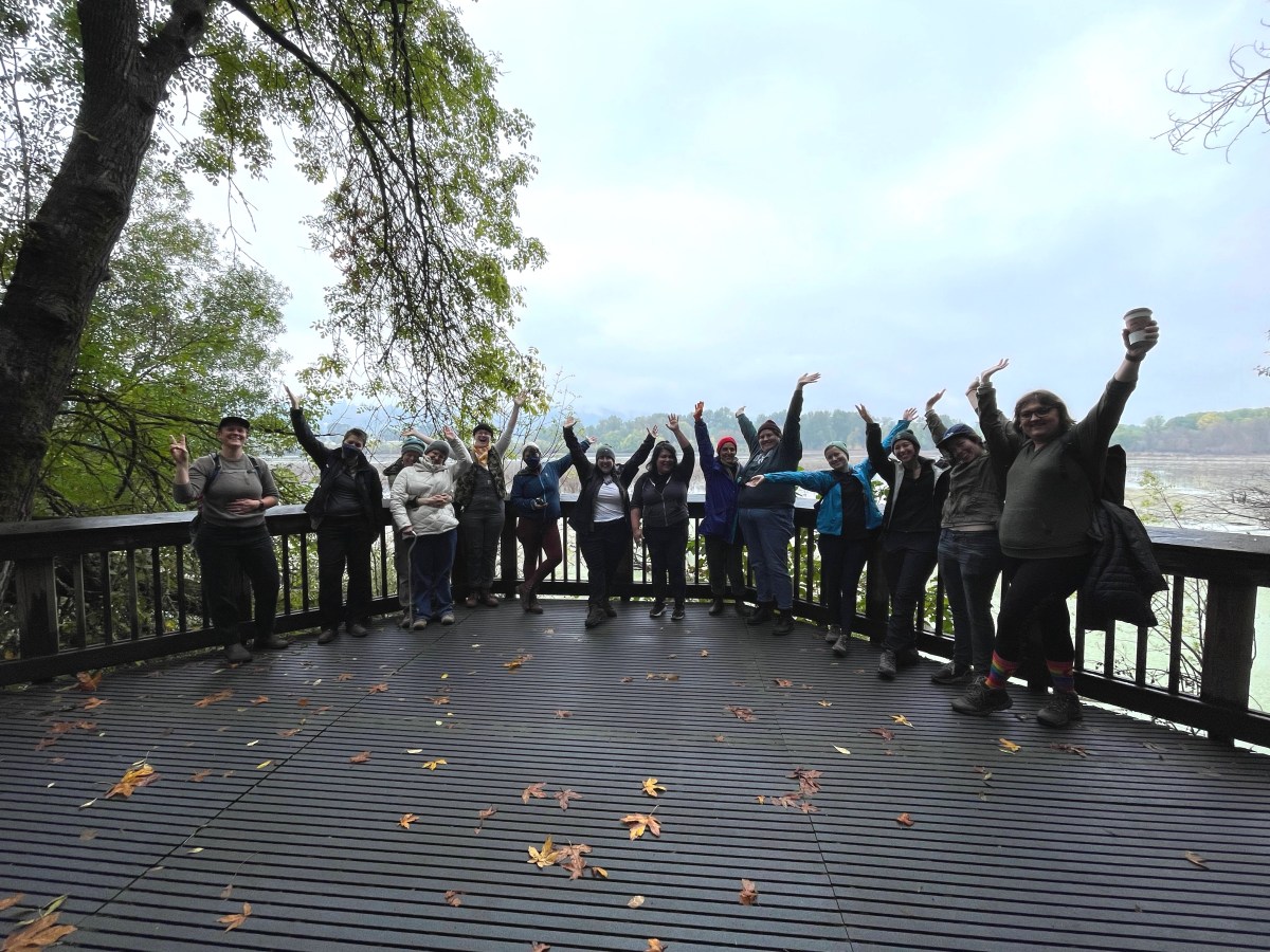 14 hikers make silly poses in front of a lake. Most throw their hands in the air. They are quite backlit.