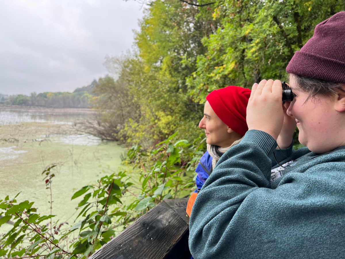 A person in a maroon hat and a teal sweatshirt holds their binoculars up to their face and smiles.