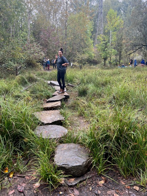 Abeni, an Autostraddle writer, holds up a peace sign with her fingers while posing on a rock path leading to the Tadpole pond. She’s tall, wearing leggings, rain boots, and an oversized sweatshirt, and her curly hair is in a messy bun. She’s grinning.