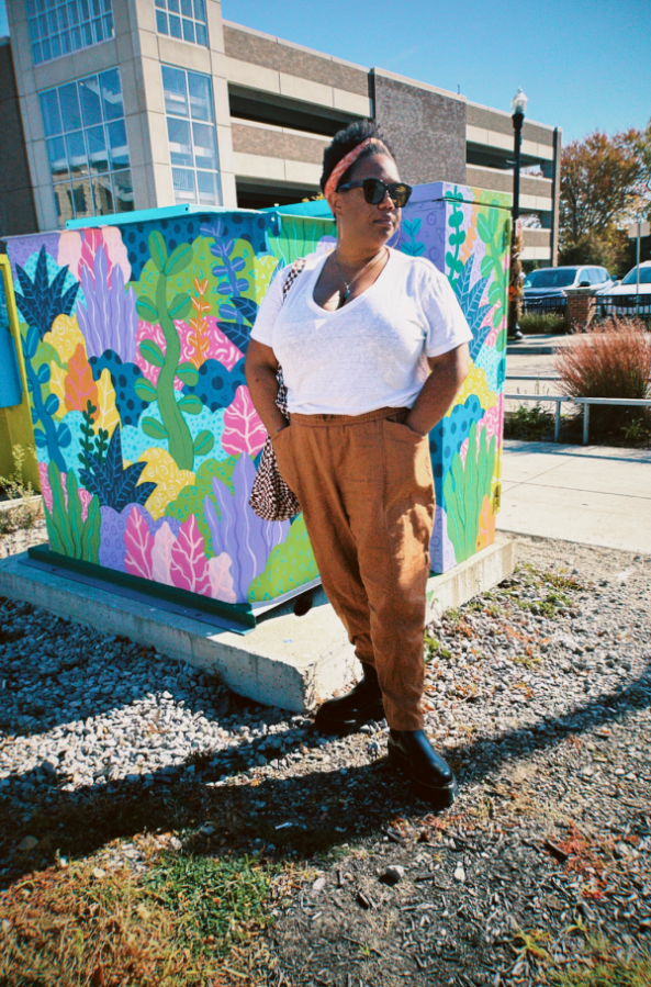 Carmen stands in a parking lot in front of a colorful spray painted structure, she has on brown pants, a white t-shirt, and her hair is bulled up with a coral headband.