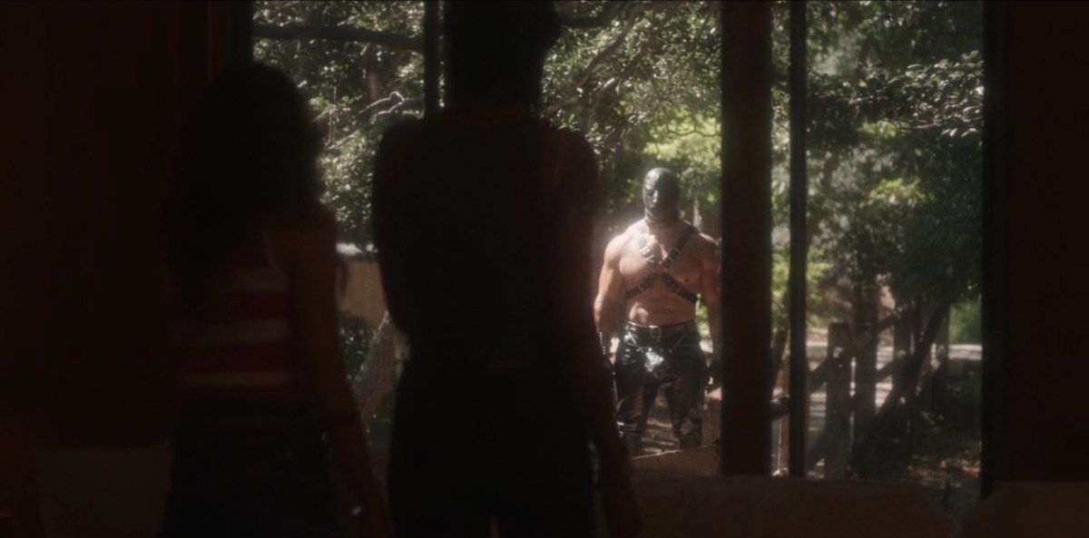 Two women backlit look out their big window at a buff man in a leather mask.