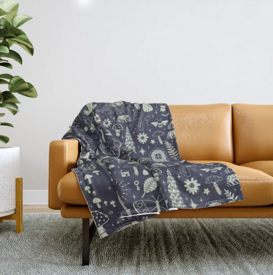 a dark blue blanket with white print draped over a light brown couch against a white wall. a tall potted plant is to the left in a white planter