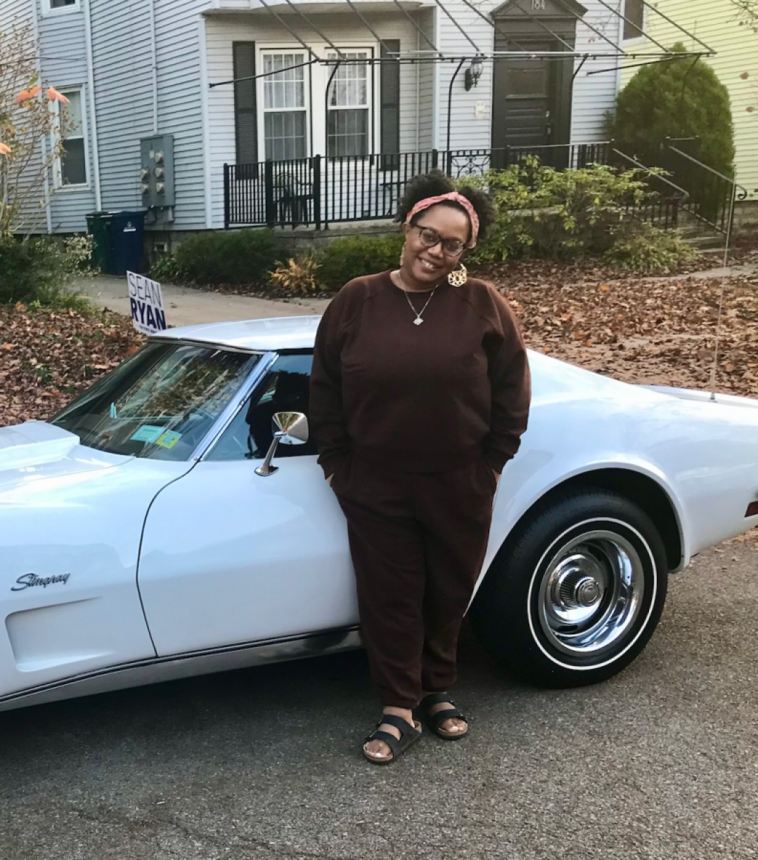 Carmen wears a deep brown sweatsuit and birkenstocks and leans against a car, smiling. Carmen is a Black woman with short black hair and glasses