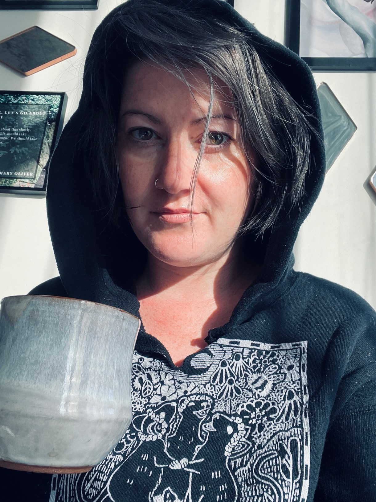 Meg is wearing a black hoodie, hood up, and drinking something from a mug. Meg is a white woman with gray hair.