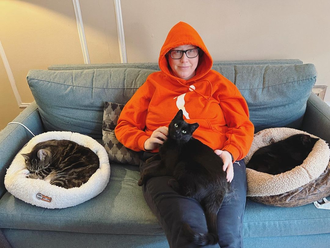 Heather is wearing. abright orange WNBA sweatshirt and cuddling with her black cat Dobbie on her couch. Heather is a soft butch white woman who wears glasses.