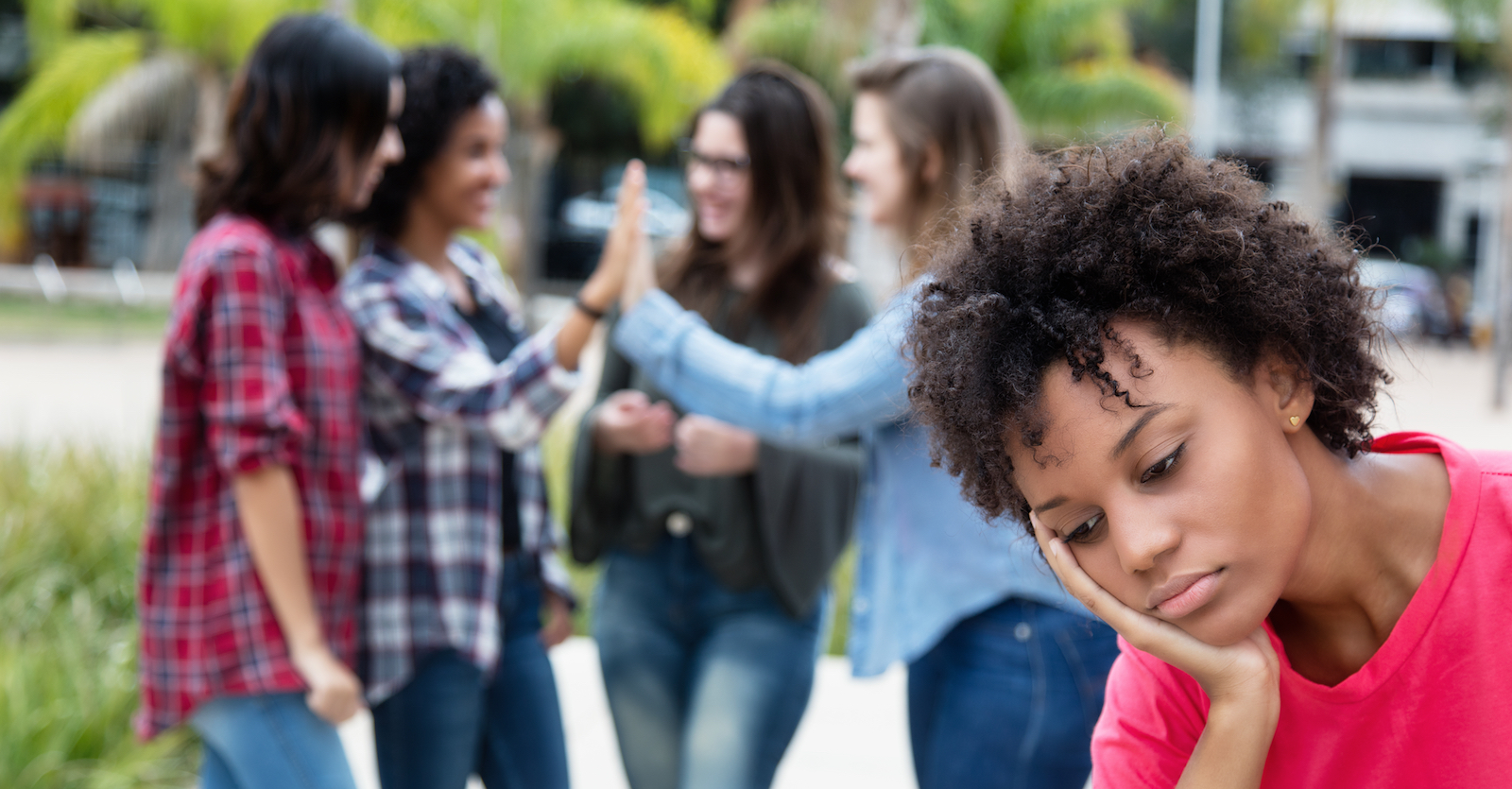 a Black woman making a sad face with her head in her hand is in the foreground, while a group of girls hang out having a nice time behind her