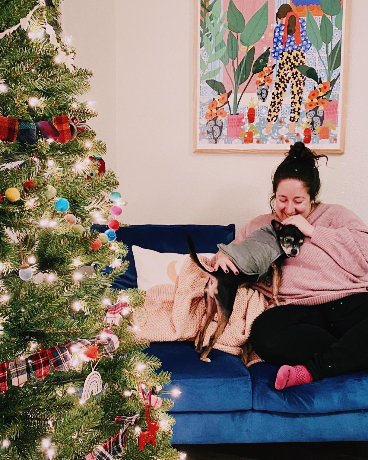 Vanessa, a white woman with brown hair, sits on a couch near a Christmas tree cuddling with her small dog