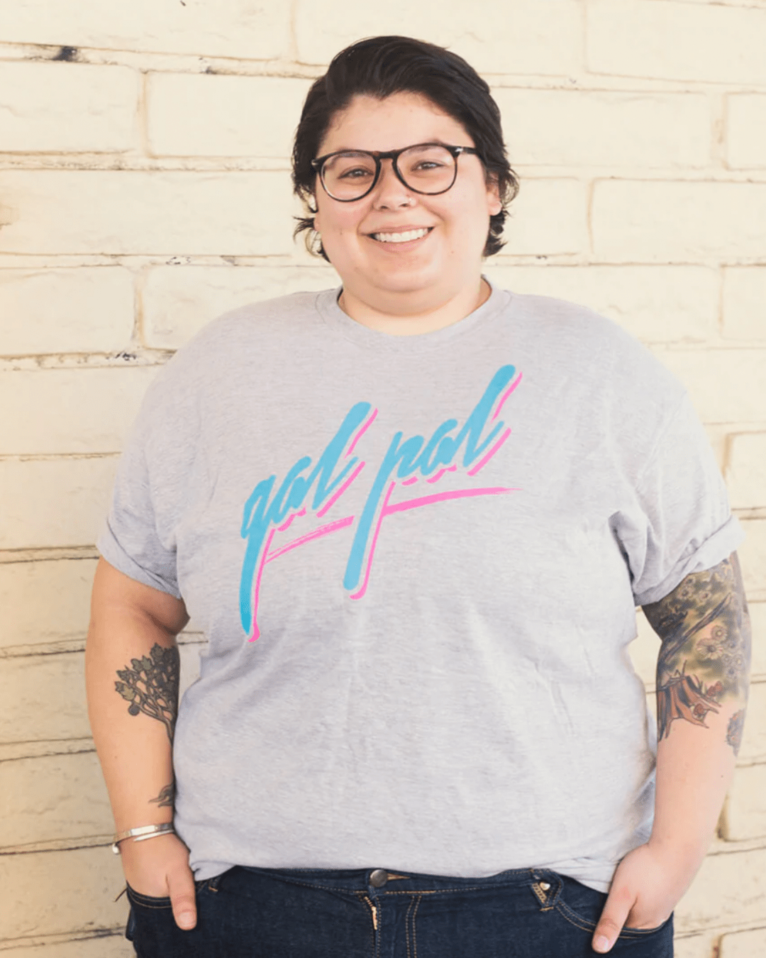 Model Chloe is wearing the Gal Pal Black Tee in size 2XL. She is 5'2" and her bra size is 38DD. The tee is light grey and has a graphic that says "gal pal" in retro 80s grafitti cursive typeface. The text is pastel blue with a pastel pink drop shadow. The text is underlined by a swipe of pink to resemble paint.