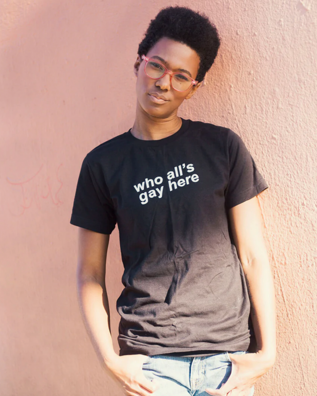 Model Sai is wearing the Who All’s Gay Here Black Tee in size S. She is 5'9" and her bra size is 34B. The tee is black with white sans-serif text that says "Who All's Gay Here." The text is askew at a diagonal in two lines.  The design is by Erin Sullivan.