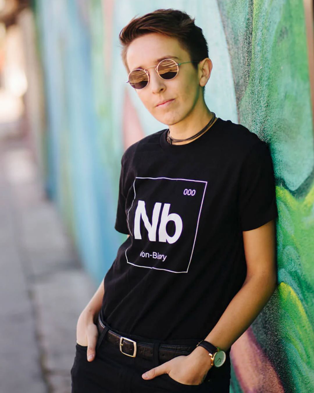 Model Ella is wearing the Non-Binary Tee in size S. They are 5'4" and their bra size is 32A. The tee is black with a white graphic resembling a unit on the periodic table. The element abbreviation is "Nb", there are numbers "000" on the top right corner, and at the bottom it says "Non-Binary."