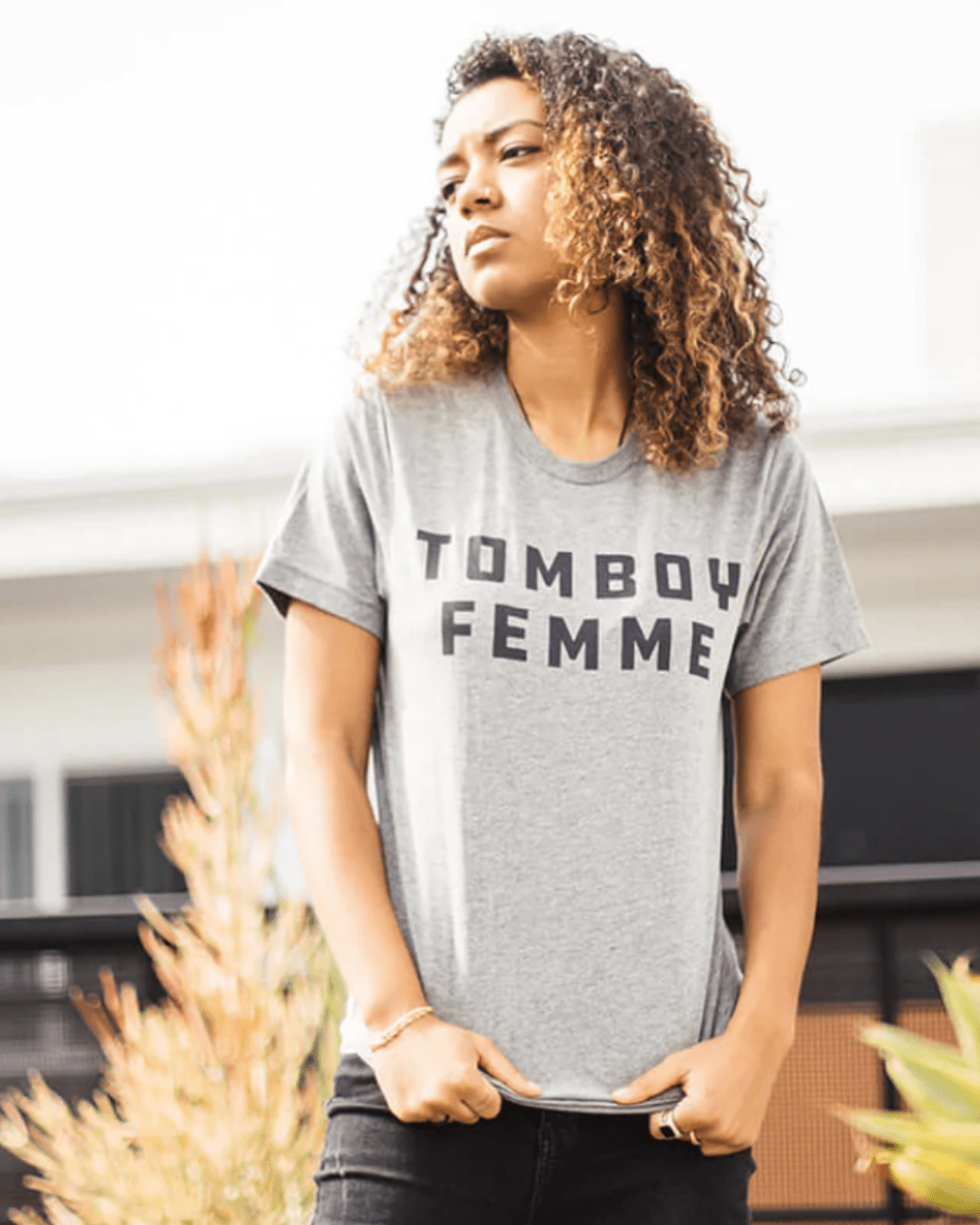 Model Alex is wearing the Tomboy Femme Tee in size S. She is 5'6" and her bra size is 34C. The tee is light grey with the words "TOMBOY FEMME' printed in all-caps in dark grey blocky type.