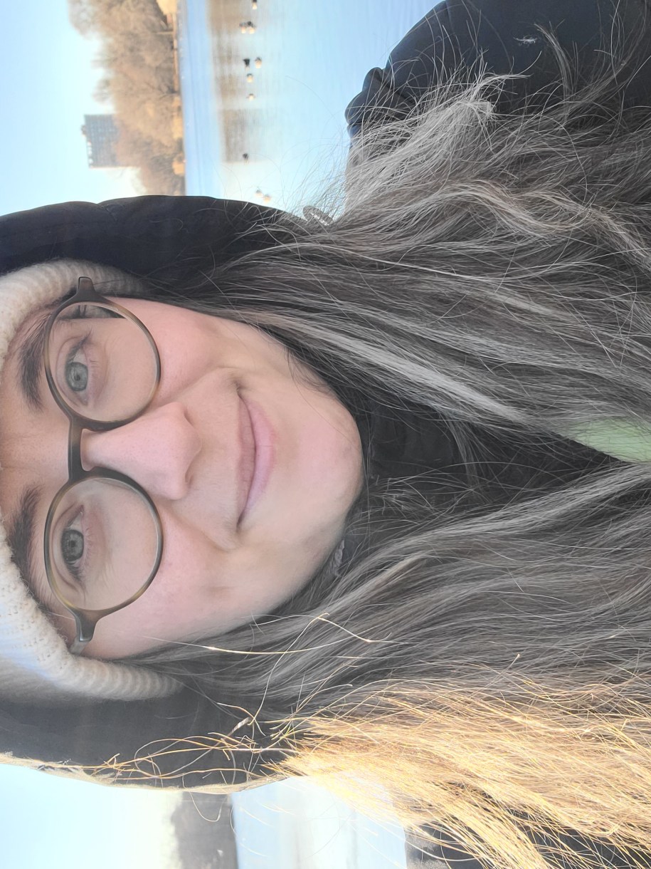 anya, a white woman with glasses and long brown hair, smiles while wearing a beanie with a smilie face on it and a jacket with a hood in front of a lake