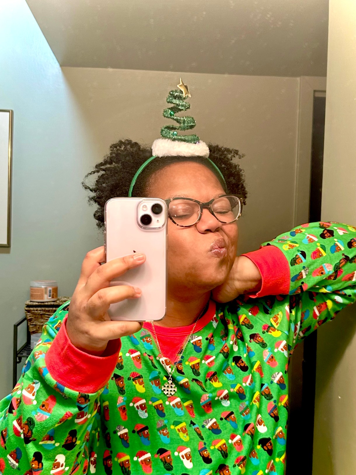 carmen a black woman with short curly hair and glasses makes a silly face while wearing red and green santa face patterned pyjamas and a christmas tree headband