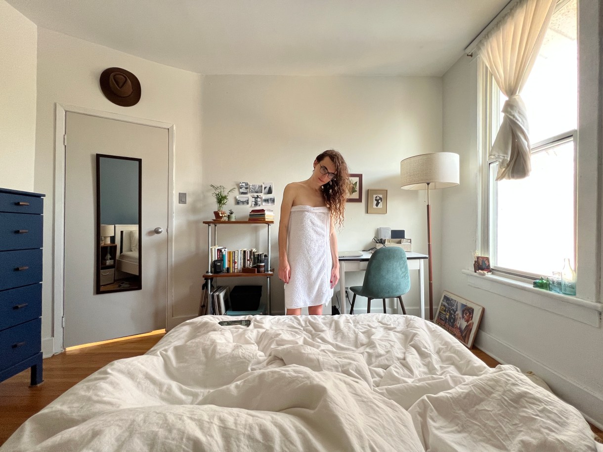 drew, a white woman wearing glasses with long curly brown hair stands far away in a room, a white towel wrapped around her, head inclined to one side