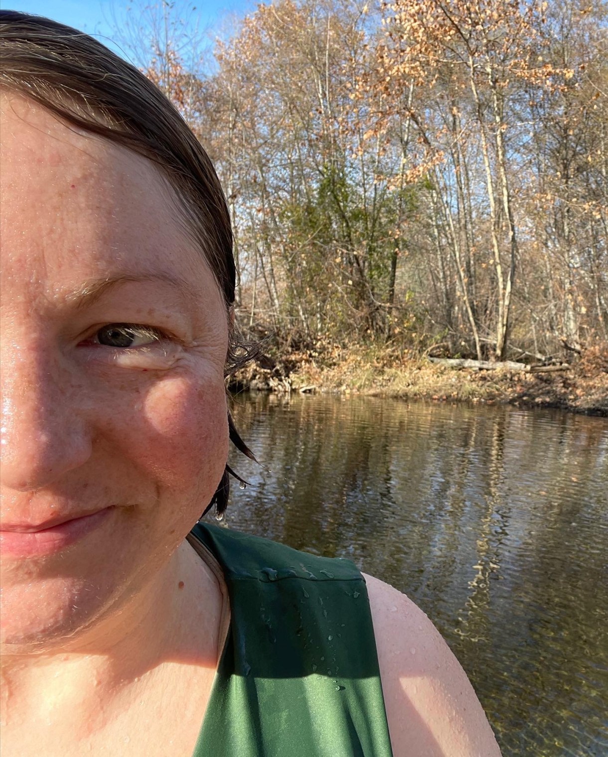 a photo of half of darcy's face, behind them a small lake. their hair is wet and they are wearing a swimsuit