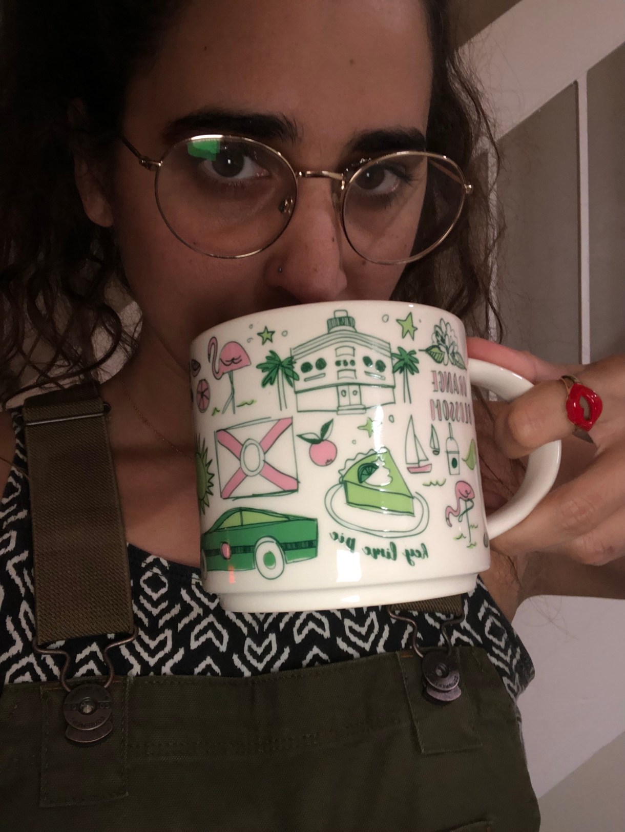 kayla, a south asian woman with glasses wears overalls and sips from a mug