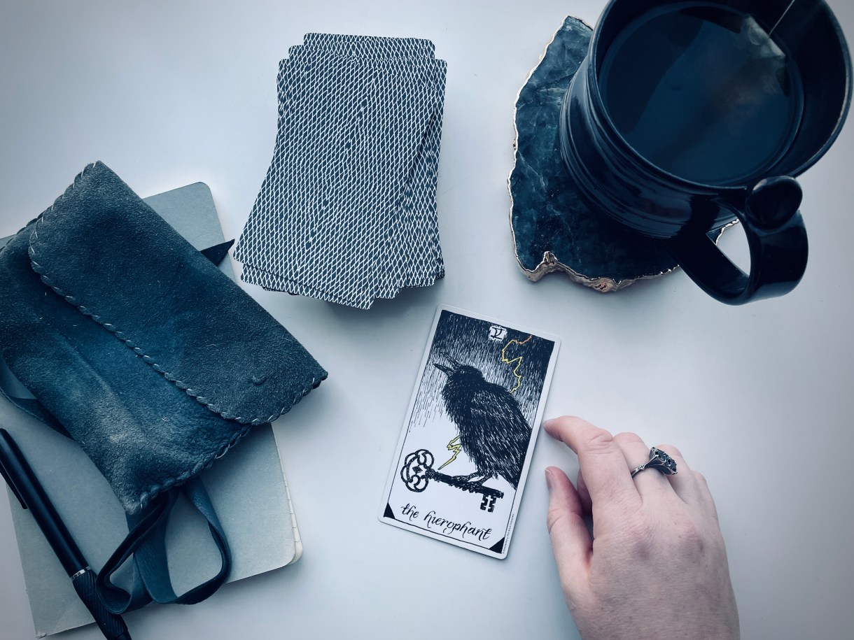 a tarot card with a crow reads "the hierophant" meg's white hand and a cup of coffee are also visible