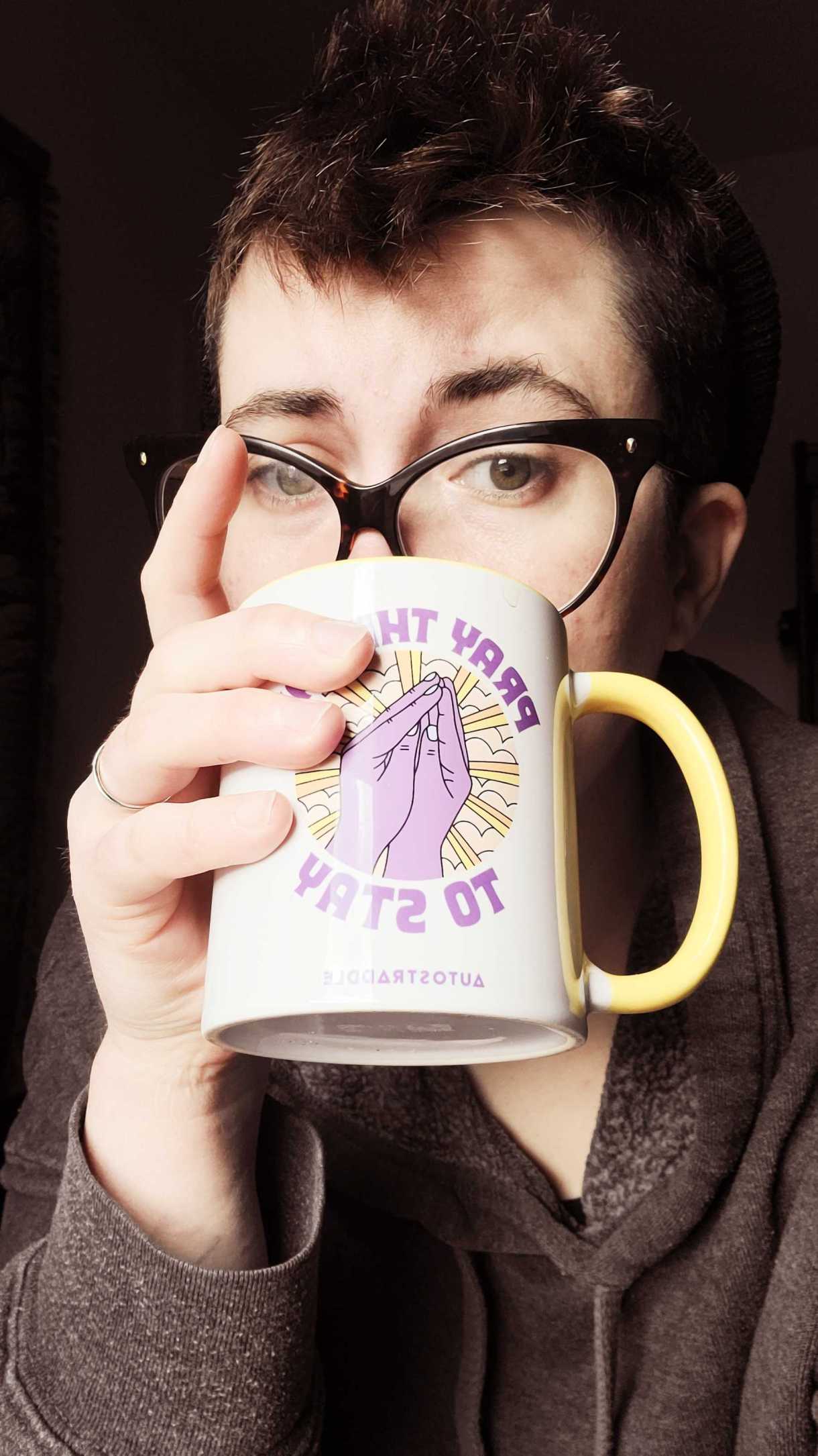 nico, a white genderqueer human with brown hair and glasses holds a "pray. the gay to stay" mug and looks over the top of it