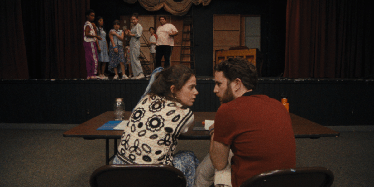 Molly Gordon and Ben Platt sit together at a directors table in a theater. Several people are on stage looking down at them. 