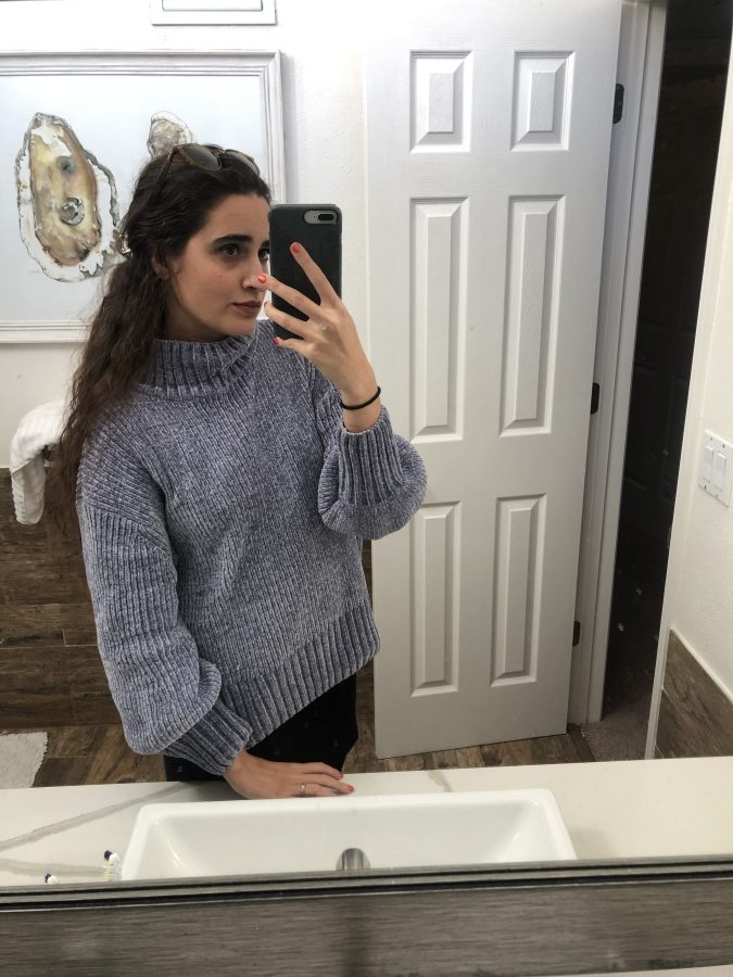 Kayla Kumari Upadhyaya wears a chunky blue-gray turtleneck sweater over a black skirt. Her hair is half up and she has sunglasses on her head.