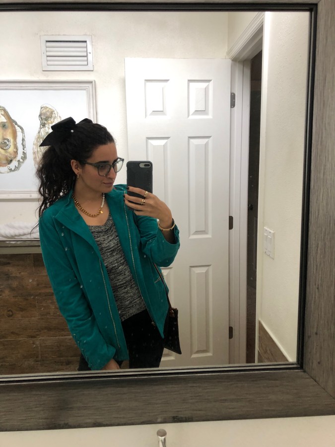 Kayla Kumari Upadhyaya wears a black and white knit blend sweater and black jeans and a teal suede motojacket. Her hair is in a ponytail with a black bow and she wears blue framed glasses and a chunky gold chain and pearl necklace.