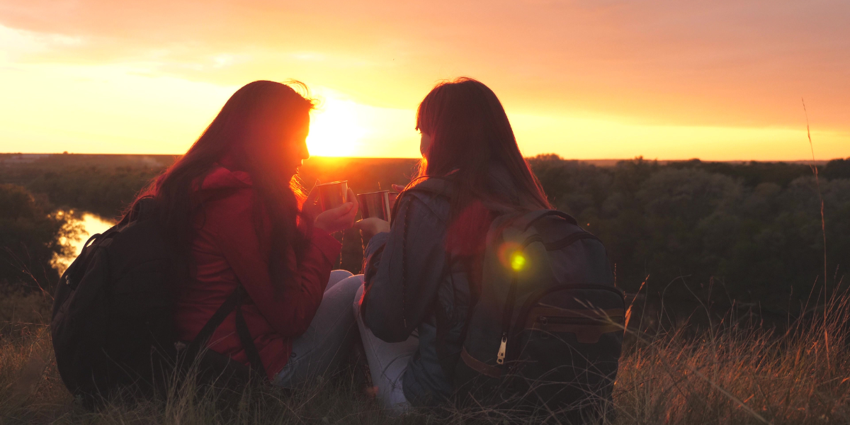 The silhouettes of two women are visible on a hill during sunset. They both have long, dark hair, wear backpacks and jeans, and hold metal cups in their hands. They sit on the ground and face each other. The woman on the left wears a red jacket and the woman on the right wears a blue jacket.