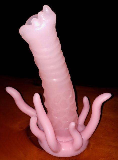 The G'lorp, a long, hollow pink dildo with ridges around the shaft and tentacles extending off of the base, sits on a red table in a dark room.