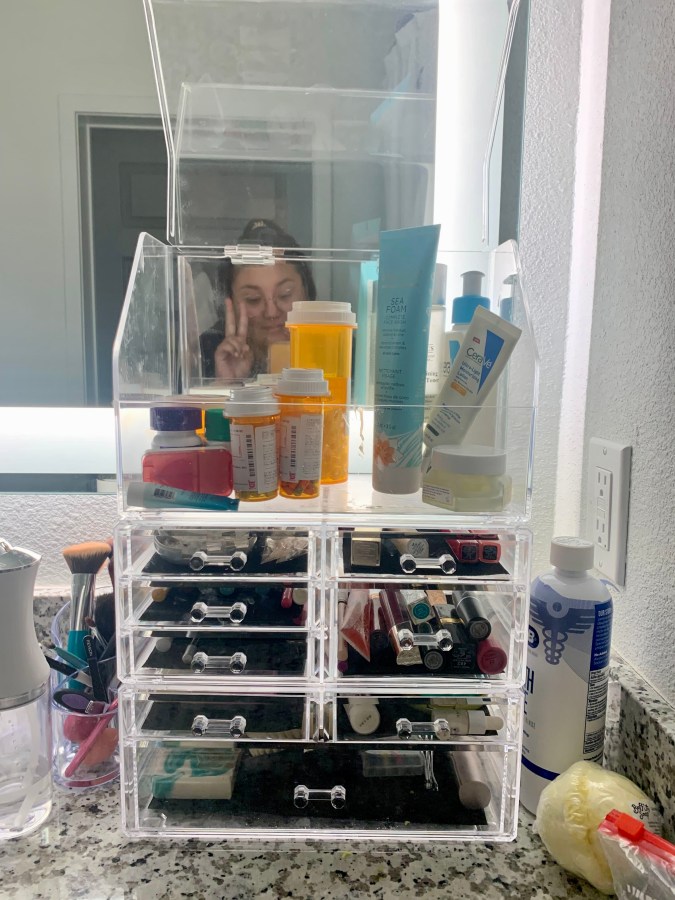 neurodiverse life hacks: a close up of clear bathroom storage containers, filled with knickknacks, stacked on top of each other. The author is making a peace sign in the back.