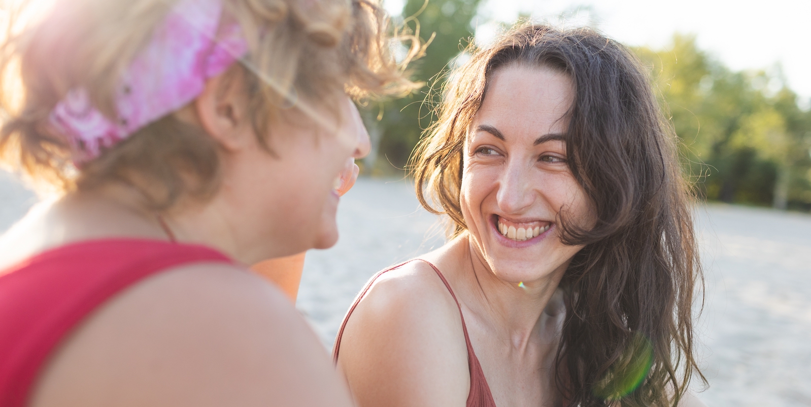 two queer people smiling at each other lovingly on a beach