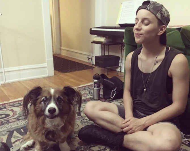 an old pic of Ro and their dear friend's dog Aragorn, who died recently. Ro is a white nonbinary human wearing a tank top, boots and backwards baseball cap. they are cross legged and smiling serenely. The dog is making a cute and silly face at the camera and his big hairy ears and a small bod and is brown and white striped