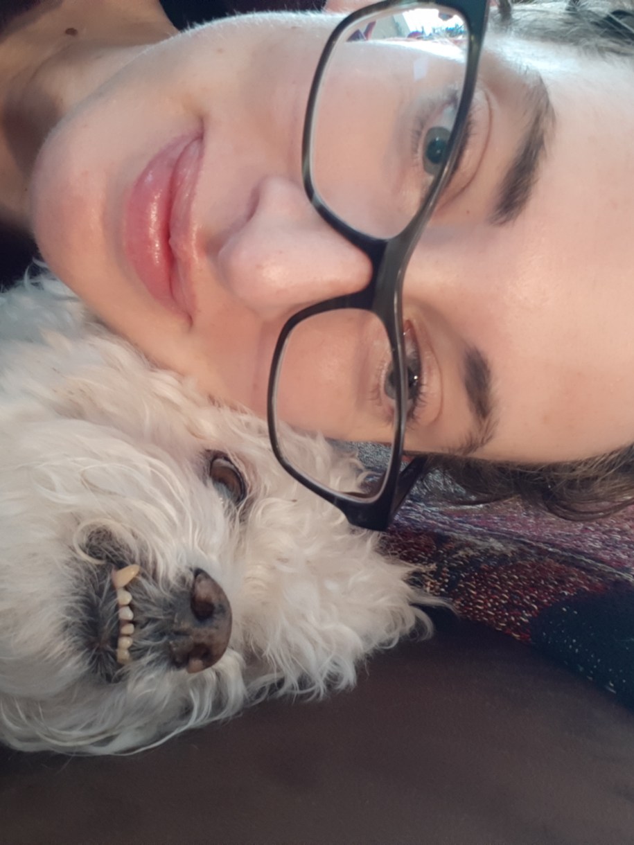 casey with an adorable floofy dog baring its tiny silly little under teeth. Casey is a white woman with glasses. this photo is just really cute. they are smushed on the couch.