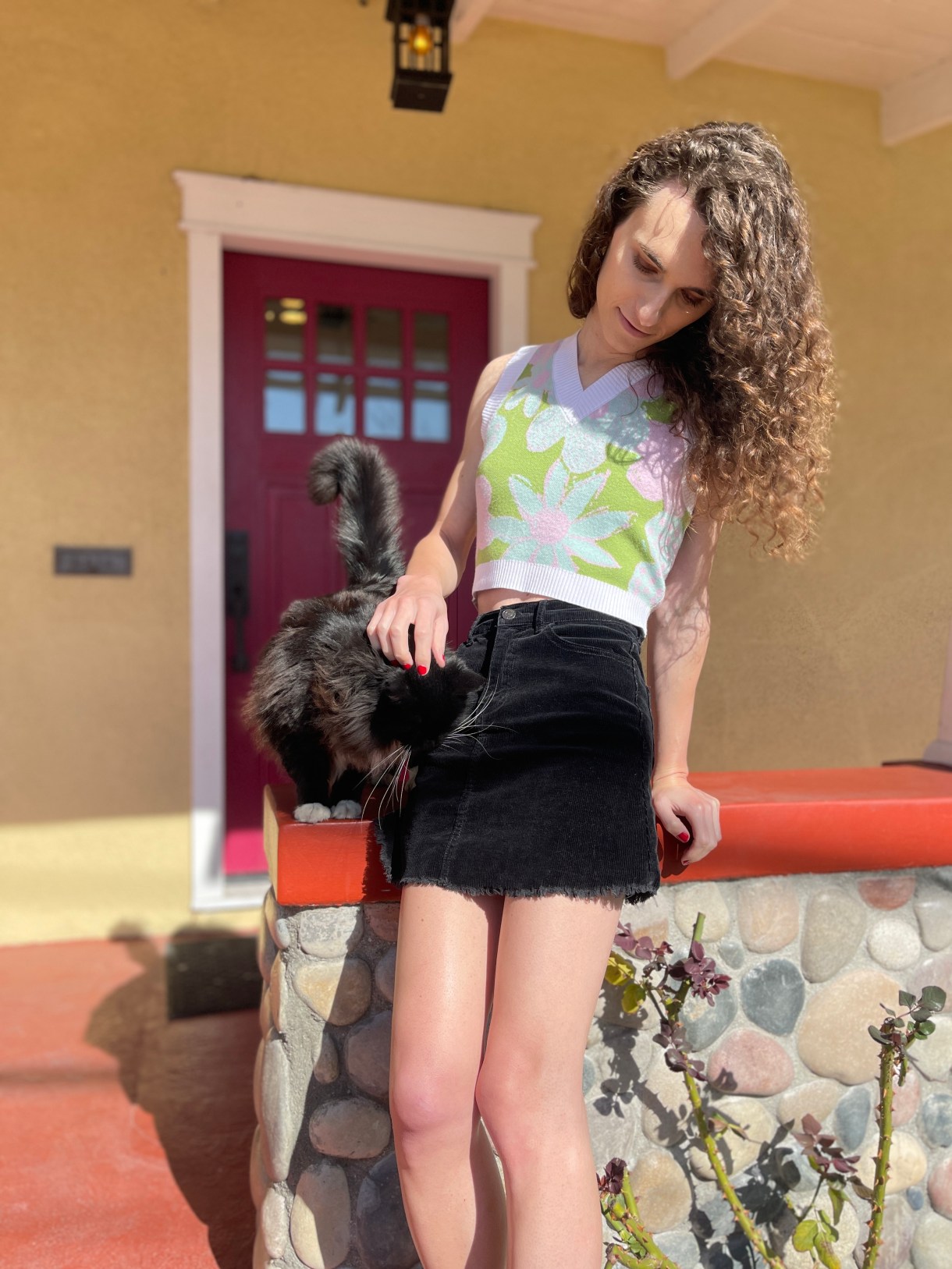 Drew leans against a wall, petting a cat, wearing a mini skirt and sleeveless knit top. She has long curly brown hair. The cat is black and floofy and butting its head into her side in order to receive more pets.