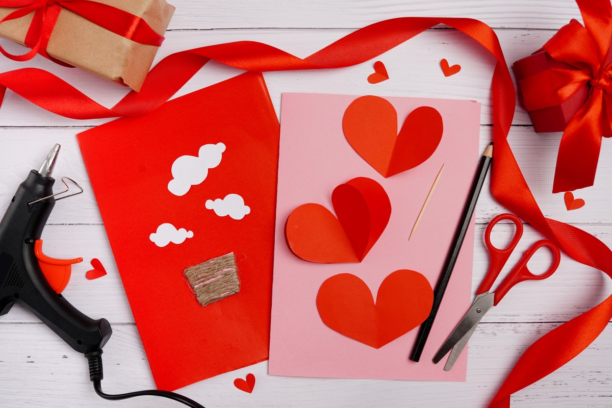 Valentine DIY supplies, including red heart cutouts, a glue gun, scissors, a pencil, ribbon, and a box