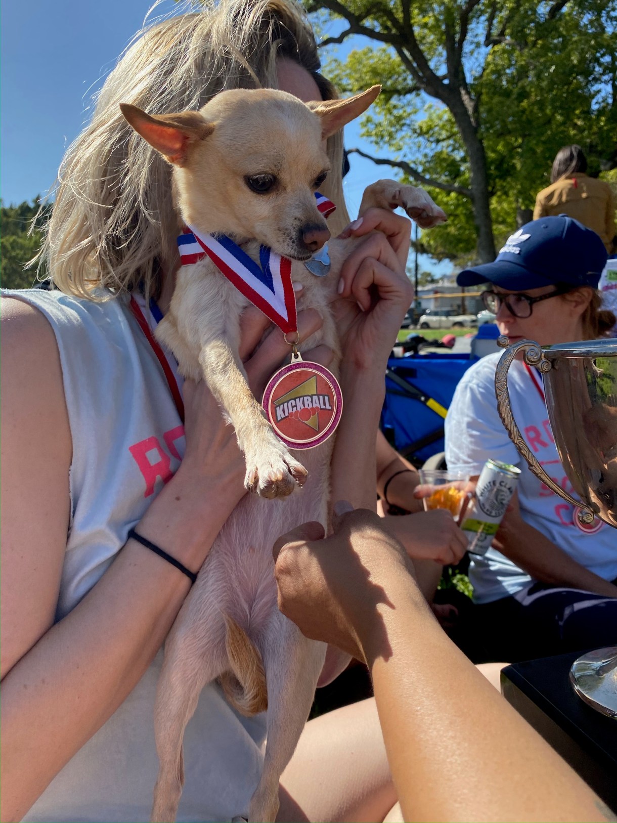 Riese holds carol the golden chihuahua in front of her face. Carol is wearing a giant sports medal and looking small and cute. Riese is a blonde white woman.